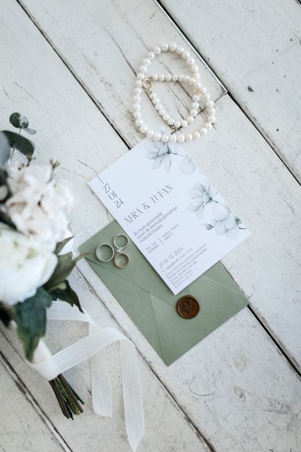 Flat lay of a wedding invitation, rings, pearls, and floral bouquet on light planks.
