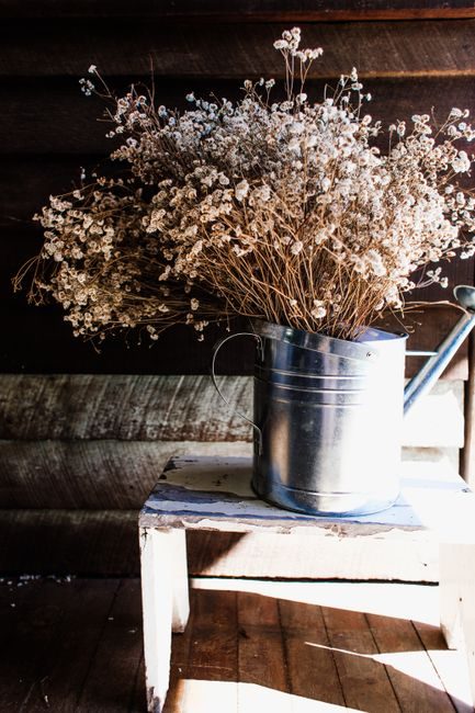 Dried flowers arranged in a metallic watering can on a white wooden stool in natural light.