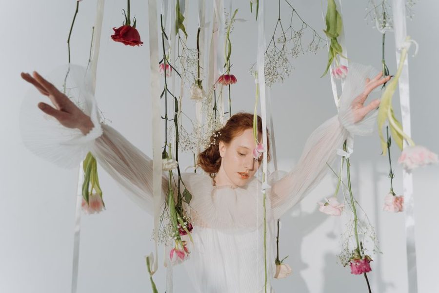 Graceful woman in white dress posing with pink and white flowers hanging around her.